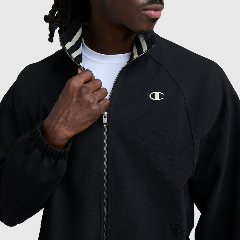 A person zips up a black Champion Gameday Full Zip Mock Neck jacket with a embroidered C logo on the left chest and striped collar lining over a white t-shirt, set against a plain light gray background.