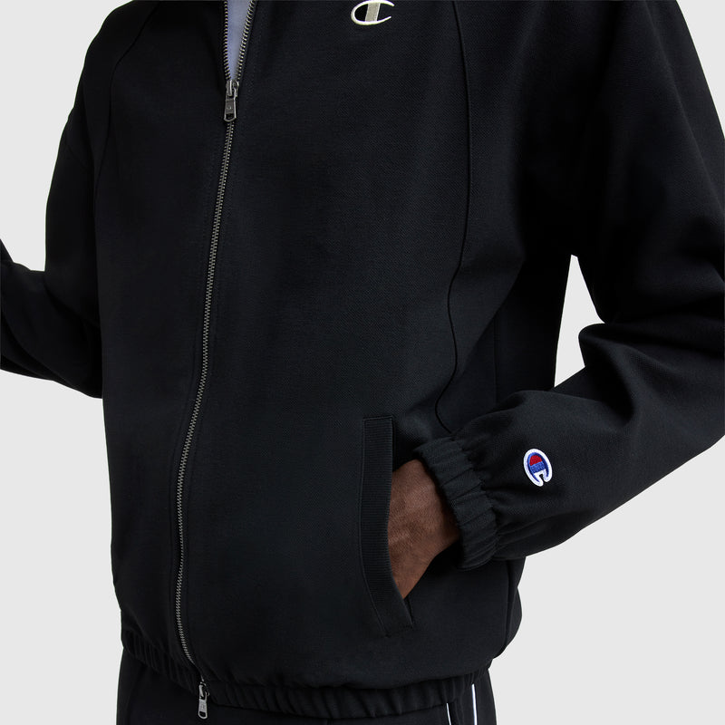 A person wears a black Champion Gameday Full Zip Mock Neck jacket with hands in pockets. The C Logo is visible on the chest and sleeve. The jacket has elastic cuffs and hem, shown against a plain white background.