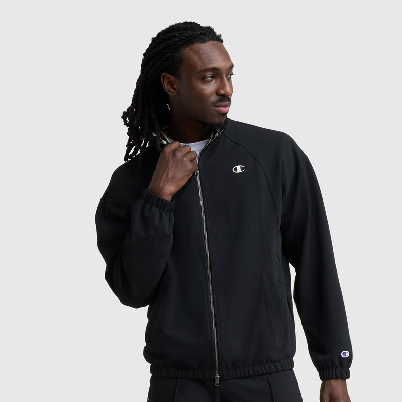 A man with long braided hair wears a black Champion Gameday Full Zip Mock Neck jacket with C logo and matching pants. He looks to the side, holding his jacket collar, against a plain light gray background.