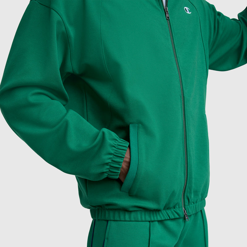 A person wears a green Champion Gameday Full Zip Mock Neck, C Logo jacket with matching green pants, one hand in their pocket, shown shoulders to hips against a light gray background.