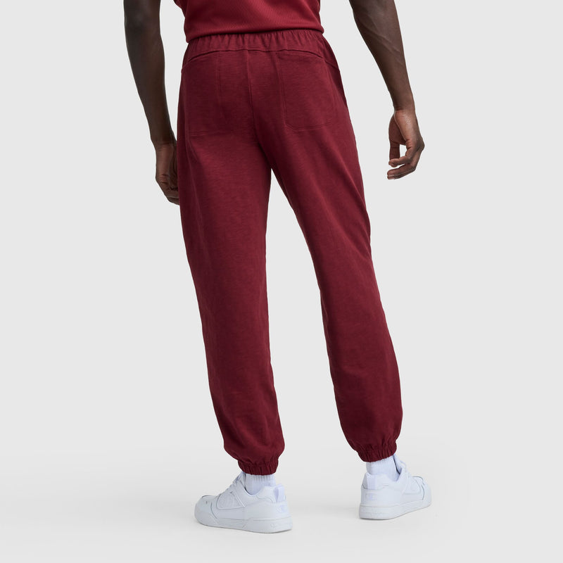 A person is seen from the back, wearing Champion’s Modern Retro Washed Pant with C Logo in maroon and white sneakers, standing against a plain light gray background with one arm relaxed at their side.