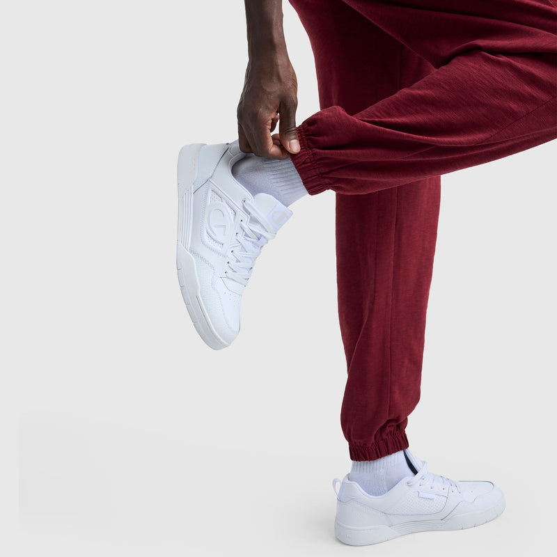 A person in Champion's Modern Retro Washed Pant, C Logo (maroon), styled with white sneakers, holds one ankle and slightly lifts their foot against a plain light background.