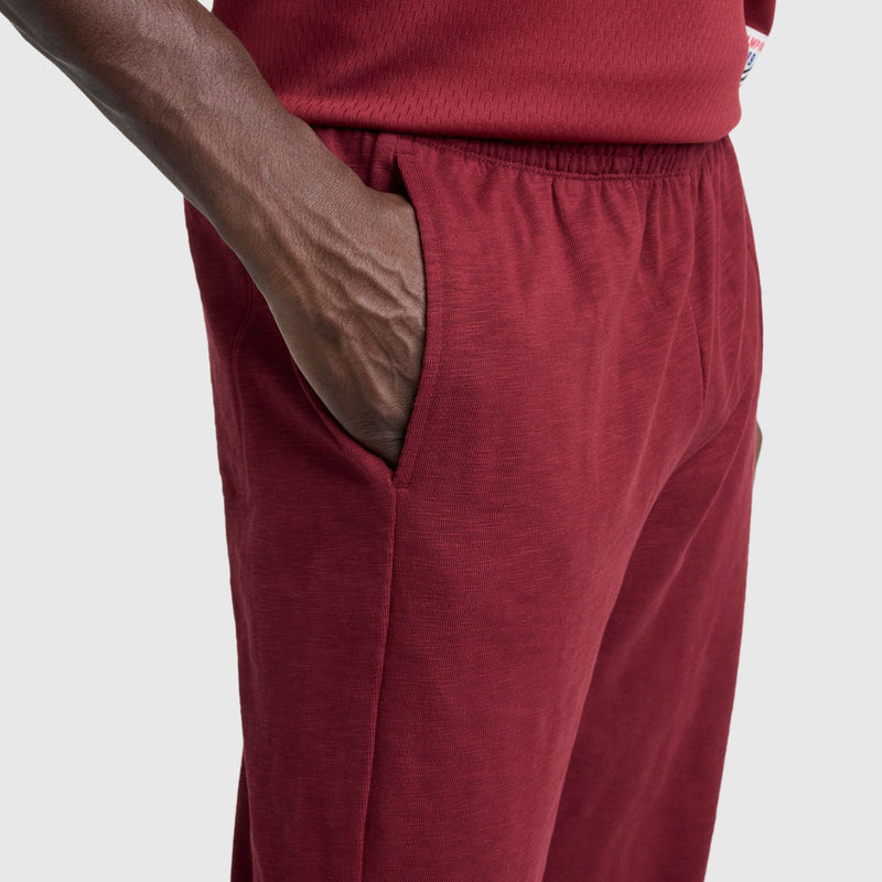 A person is shown from the waist down against a plain light gray background wearing Champion’s Modern Retro Washed Pant, C Logo in red, with their right hand in their pocket.