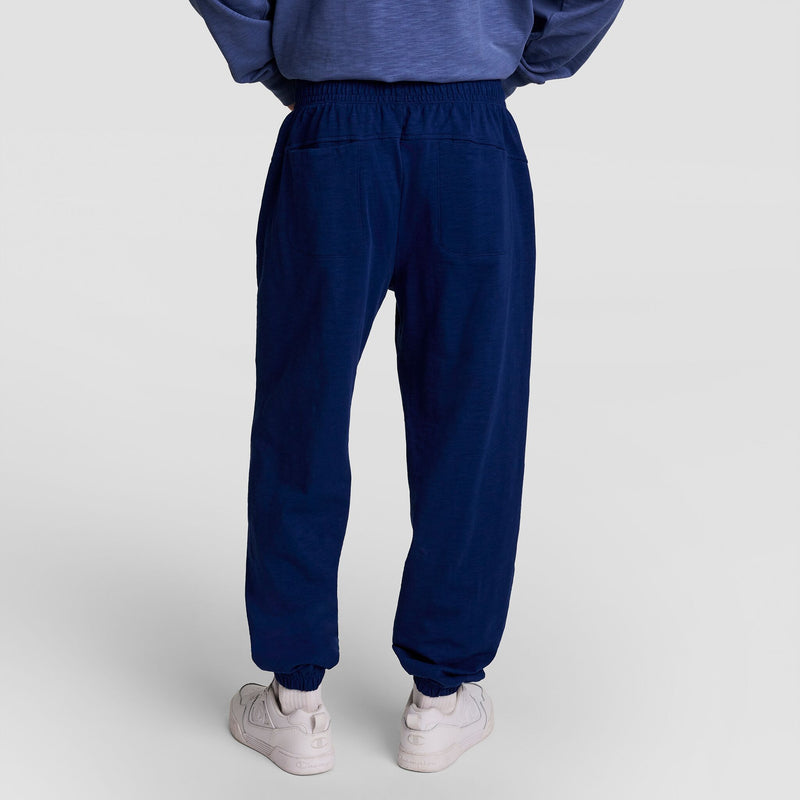 A person wears Champion’s Modern Retro Washed Pant, C Logo in navy blue heavy textured slub cotton with white sneakers, shown from the back against a plain light background. Only the lower half is visible, featuring subtle Champion embroidery.