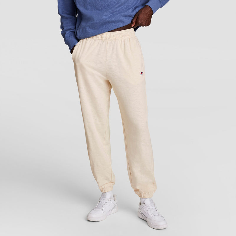 A person wearing Champion’s Modern Retro Washed Pant with the C Logo, a blue long-sleeve shirt, and white sneakers stands with one hand in their pocket against a plain light background.