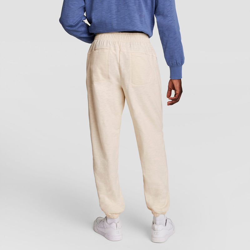 Someone stands with their back to the camera against a plain light background, wearing Champion’s Modern Retro Washed Pant, C Logo joggers, a blue long-sleeve shirt, and white sneakers.