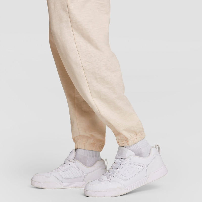 A person wears Champion’s Modern Retro Washed Pant, C Logo in light beige, teamed with white crew socks and white sneakers, standing against a plain light gray background; only their lower legs and feet are visible.