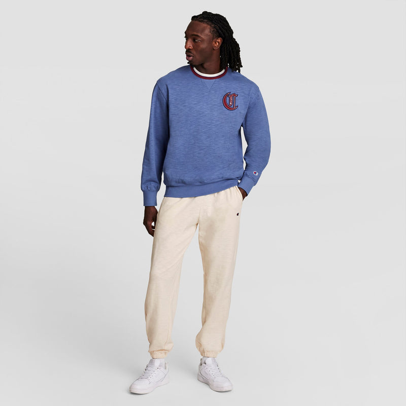 A man stands against a plain background wearing a blue Champion crewneck sweatshirt, Modern Retro Washed Pant joggers with the C Logo by Champion in light beige, and white sneakers. He looks left with one hand in his pocket.