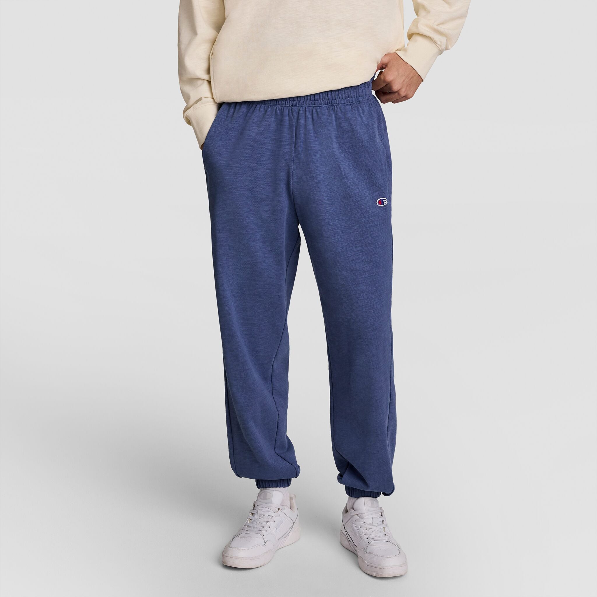 A person wearing a cream sweatshirt, Champion Modern Retro Washed Pant with C Logo in blue, and white sneakers stands with hands in pockets against a plain light background.