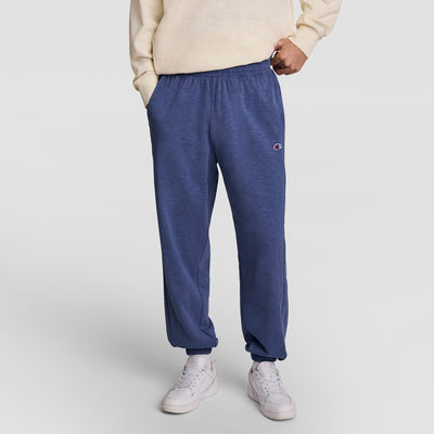 A person wearing a cream sweatshirt, Champion Modern Retro Washed Pant with C Logo in blue, and white sneakers stands with hands in pockets against a plain light background.