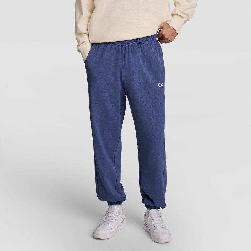 A person wearing a cream sweatshirt, Champion Modern Retro Washed Pant with C Logo in blue, and white sneakers stands with hands in pockets against a plain light background.