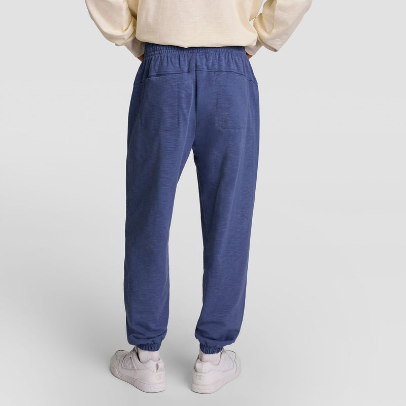 A person is shown from the back wearing a cream sweatshirt, Champion’s Modern Retro Washed Pant, C Logo in blue, and white sneakers, standing on a plain light background.