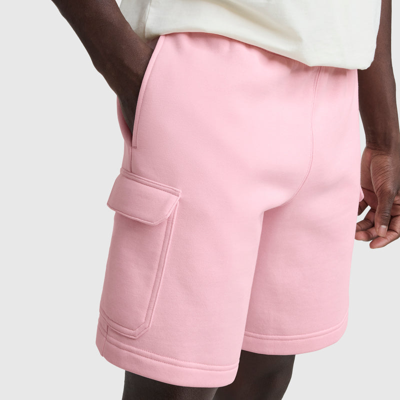 A person wears Champion's 8" Fleece Cargo Short with the C Logo in light pink, made from a cotton-blend fleece with a large side pocket, paired with a white shirt, hand in pocket, set against a plain light gray background.