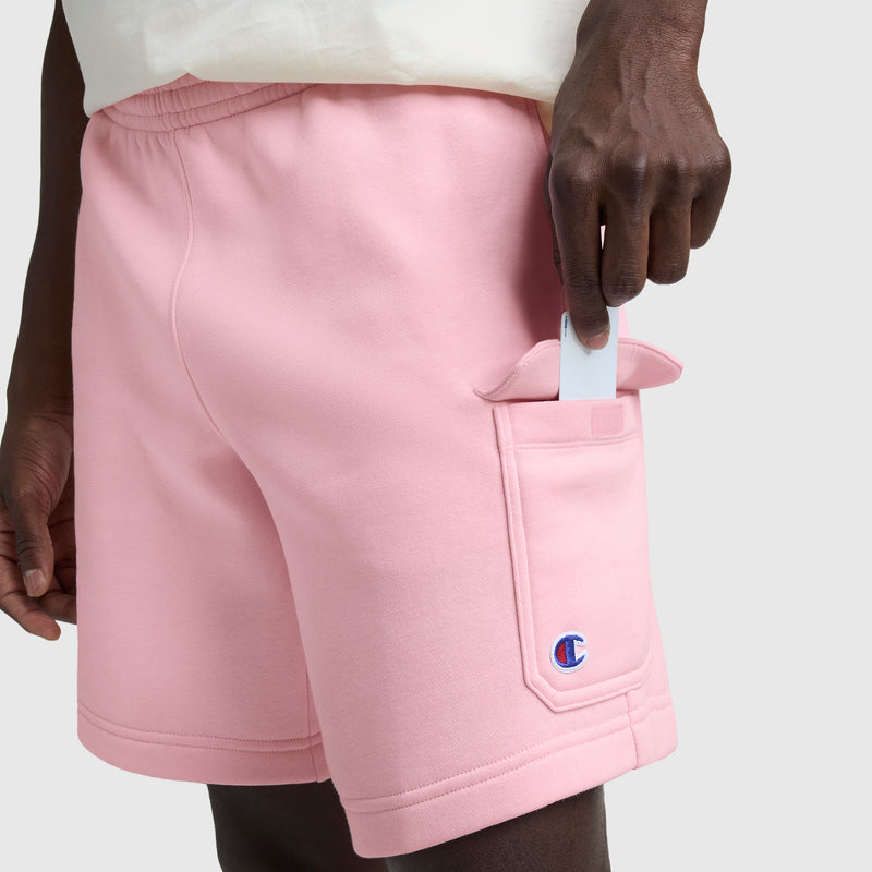 A person wearing Champion 8" Fleece Cargo Shorts with the C Logo in light pink is shown putting a white smartphone into the back pocket, highlighting the cotton-blend fleece against a plain light gray background.
