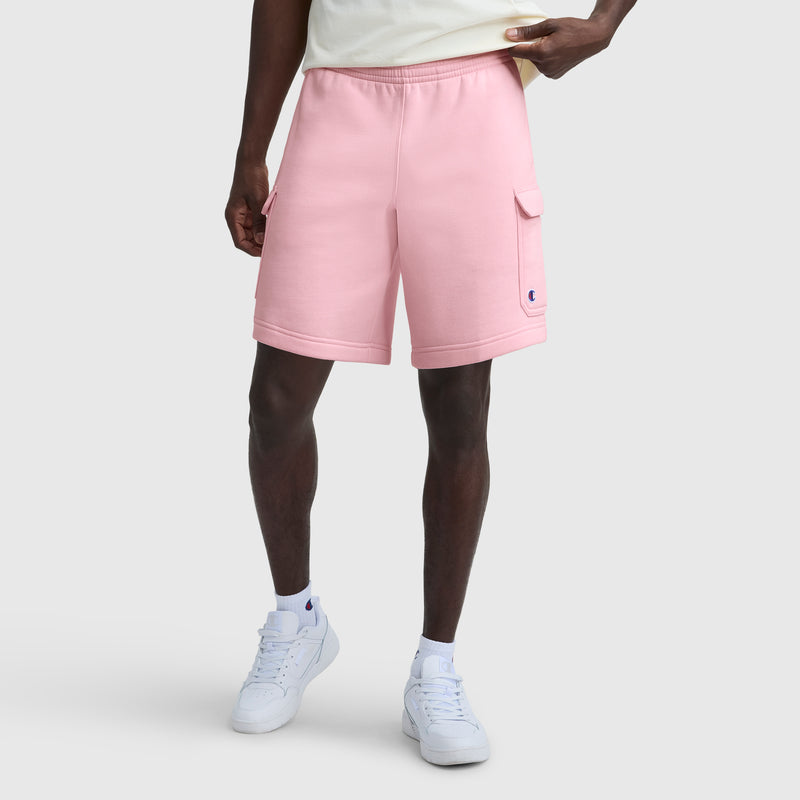 A person is shown from the waist down wearing Champion’s 8" Fleece Cargo Short with C Logo in light pink, paired with a light t-shirt, white socks, and white sneakers against a plain gray background.