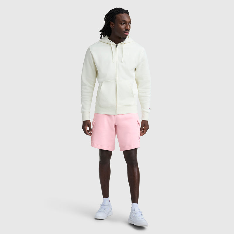 A man stands facing slightly left, wearing a cream cotton-blend fleece zip-up hoodie, Champion 8" Fleece Cargo Short with C Logo in light pink, and white sneakers against a plain light gray background.