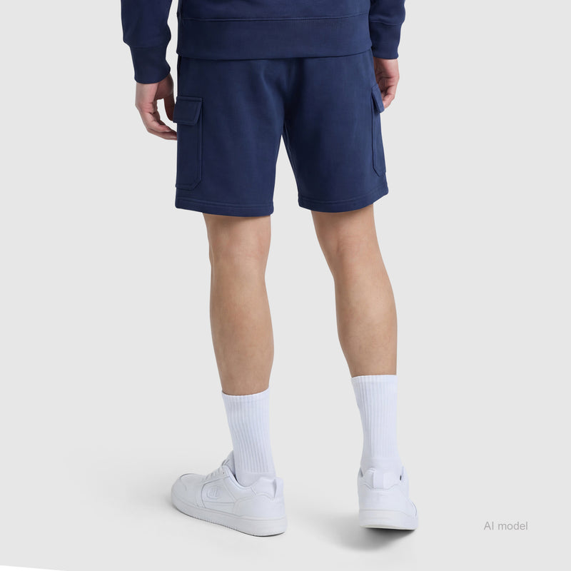 Standing with their back to the camera, the person is wearing Champion’s 8" Fleece Cargo Short with the C Logo in navy blue cotton-blend fleece, paired with a matching sweatshirt, white crew socks, and white sneakers against a plain light background.