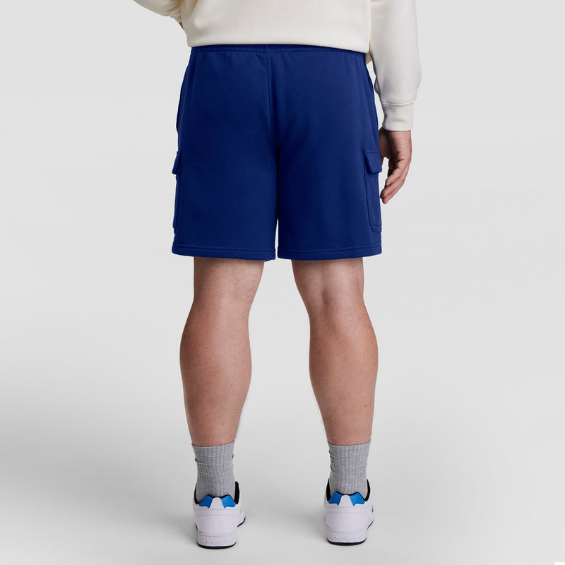 Shown from the back against a plain light background, the person wears Champion 8" Fleece Cargo Shorts (C Logo - Big & Tall), paired with gray socks, white sneakers with blue accents, and a cream sweatshirt. Cotton-blend fleece ensures comfort.
