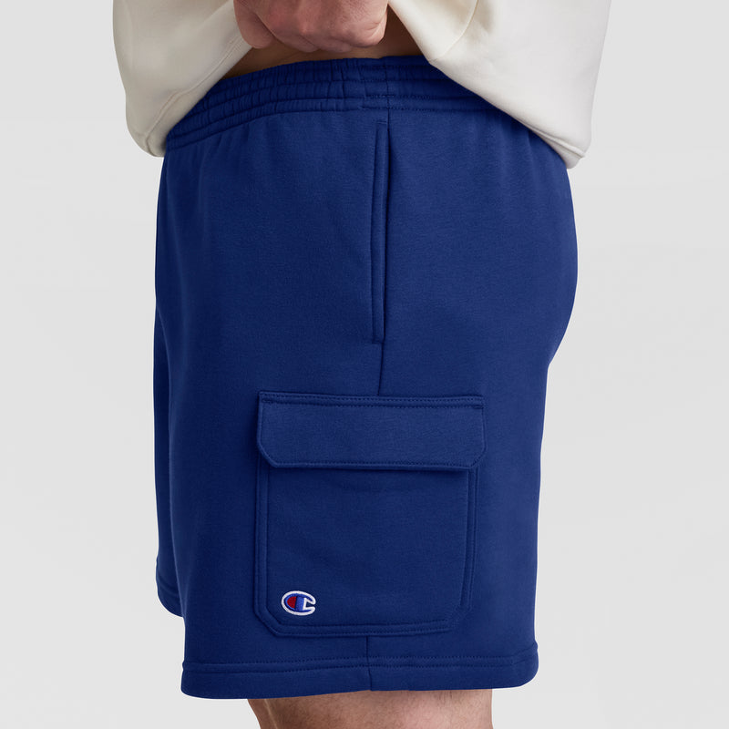 A person wears Champion’s Big & Tall 8" Fleece Cargo Short in royal blue, featuring cotton-blend fleece, an elastic waistband, side flap pocket, and a small C logo. The upper part of a light shirt is partially visible.