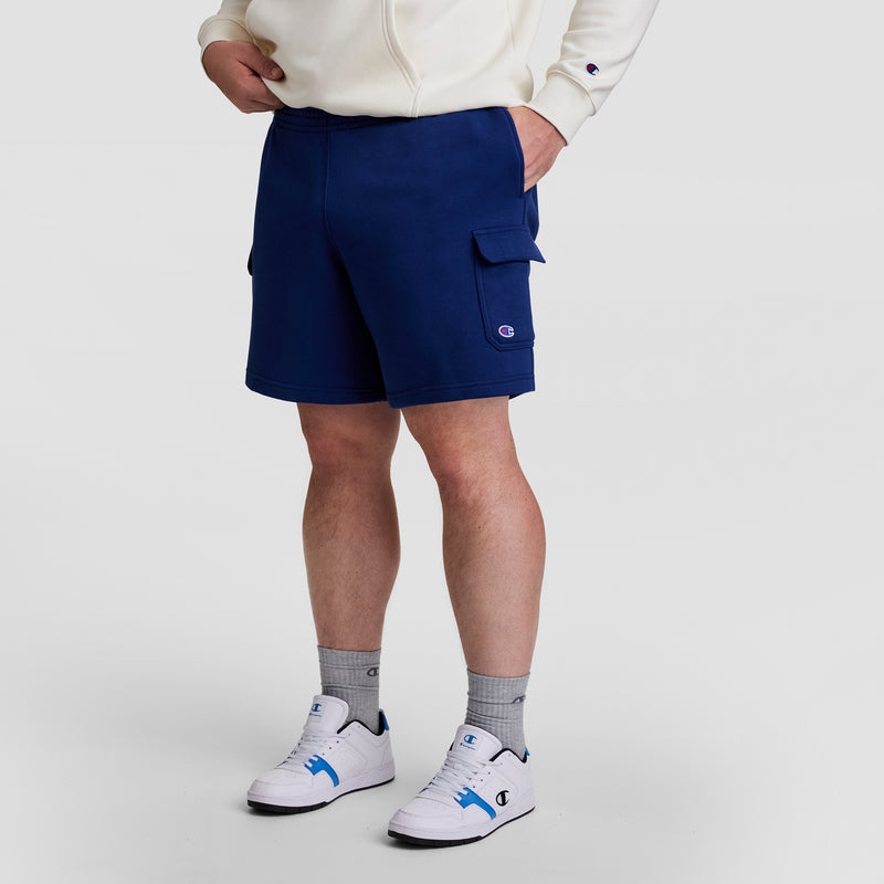 A person wearing a white hoodie, Champion 8" Fleece Cargo Short, C Logo - Big & Tall in blue, gray Champion socks, and white Champion sneakers with blue accents stands against a plain light background.