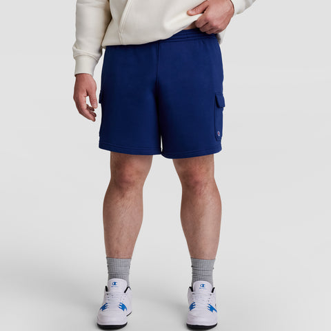 A person in a cream hoodie, Champion 8" Fleece Cargo Short C Logo - Big & Tall (blue cotton-blend), gray socks, and white sneakers with blue accents stands against a plain light background. Their face is not visible.