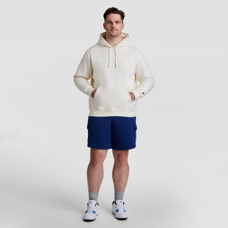 A man stands facing forward in a cream-colored hoodie, Champion 8" Fleece Cargo Short with C Logo (Big & Tall) in navy, gray socks, and white sneakers, against a plain light gray background.
