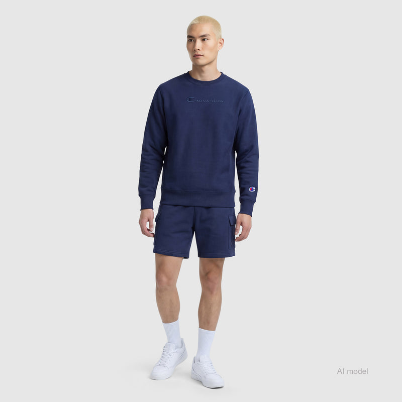 A person with short blond hair wears a navy blue Champion sweatshirt and matching Champion 8" Fleece Cargo Shorts with the C Logo, paired with white socks and white sneakers, standing against a plain light gray background.