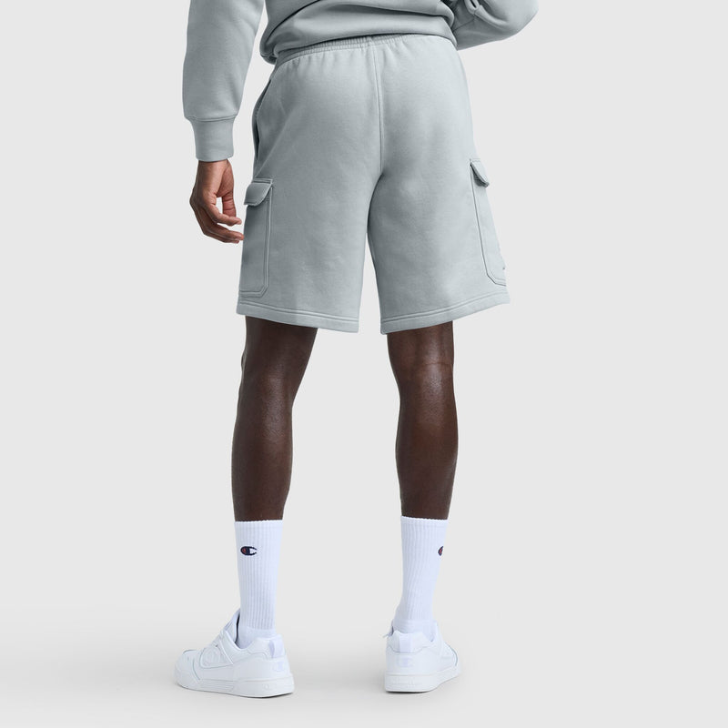 A person stands facing away, wearing Champion 8" Fleece Cargo Shorts with C Logo in light gray and a matching sweatshirt, plus white logo socks and white sneakers, against a plain background.