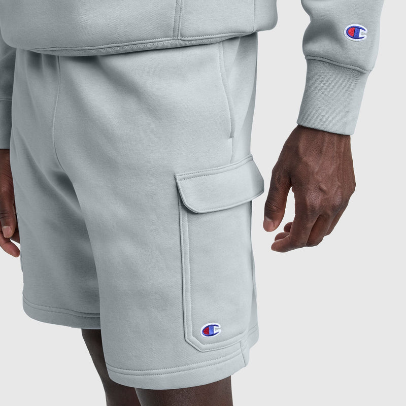 Someone wears Champion's 8" Fleece Cargo Short in light gray, featuring the C logo on the pocket, paired with a matching sweatshirt showing the sleeve logo. Their right hand rests relaxed by their side.