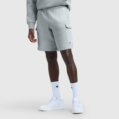 A person wears Champion's 8" Fleece Cargo Shorts with a C Logo in light gray, paired with a matching sweatshirt, white Champion socks, and white sneakers against a gray background. The photo is chest-up and shows relaxed hands.