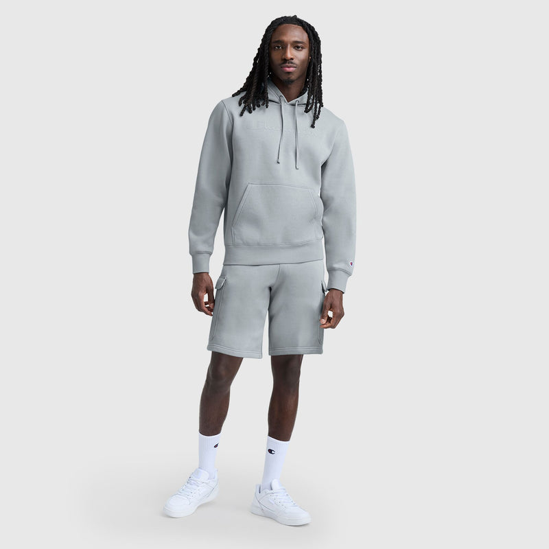 A man with long dreadlocks stands against a plain light gray background wearing a light gray hoodie, Champion 8" Fleece Cargo Short with C Logo, white socks, and white sneakers.