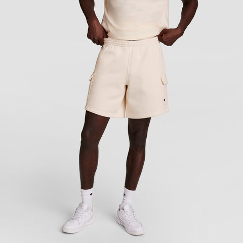 A person stands against a plain, light background wearing cream Champion 8" Fleece Cargo Shorts (C Logo), a matching cream t-shirt, white crew socks, and white sneakers, with hands resting on their hips.
