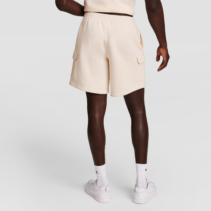 A person stands facing away, wearing Champion 8" Fleece Cargo Shorts with C Logo in light beige, a matching beige top, white socks, and white sneakers against a plain white background.