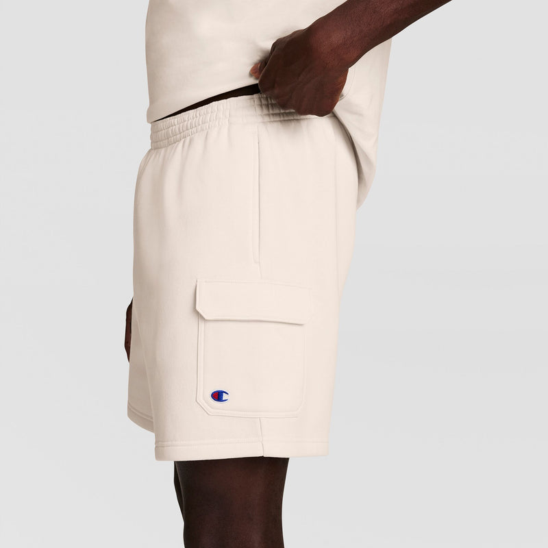 Wearing Champion's 8" Fleece Cargo Short with C Logo in off-white, featuring soft cotton-blend fleece, an elastic waistband, and a large side pocket. Only the lower torso and upper thighs are shown against a plain background.