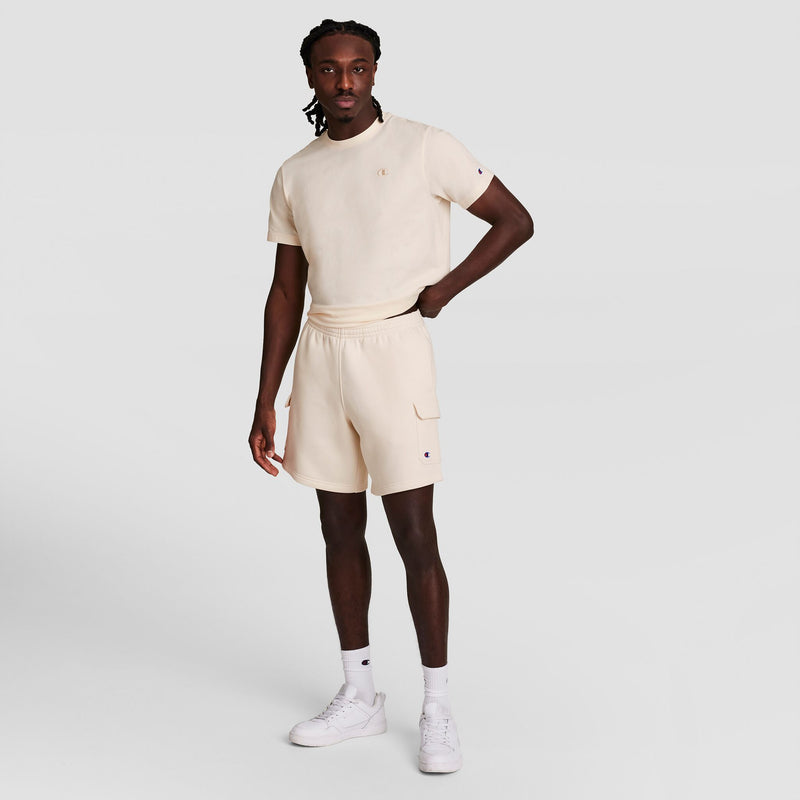 A man stands confidently in Champion's 8" Fleece Cargo Short with a C logo, styled as part of a matching cream cotton-blend fleece set, white socks, and white sneakers, against a plain light gray background.
