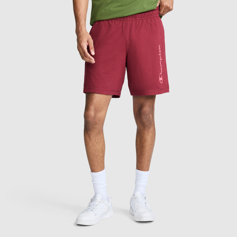 A person wearing a green shirt, maroon Champion 7" Jersey Shorts with Script Logo, white crew socks, and white sneakers stands against a plain light background.