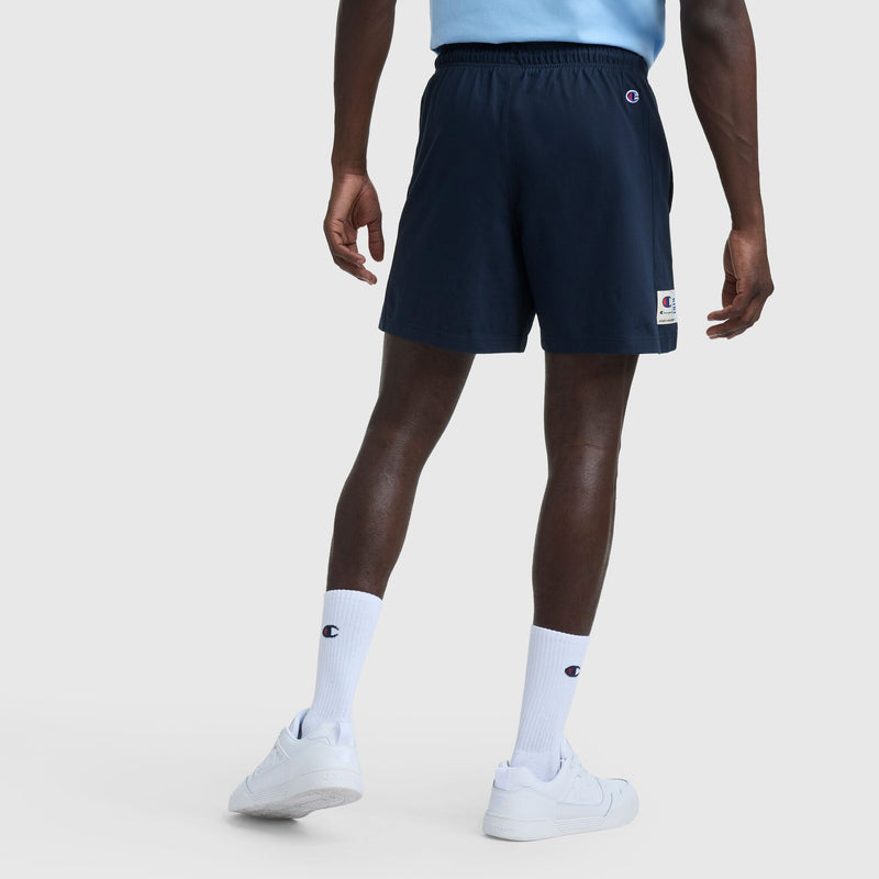 A person faces away, wearing a light blue heritage cotton jersey shirt, Champion 7″ Jersey Shorts, Script Logo in navy, white crew socks with Champion script, and white sneakers against a plain light background.