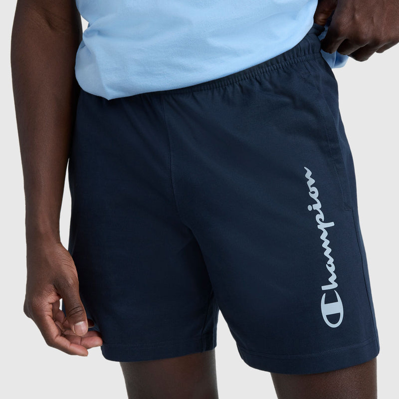A person wearing a light blue shirt and Champion 7″ Jersey Shorts, Script Logo, featuring the vertical script printed on the left leg.