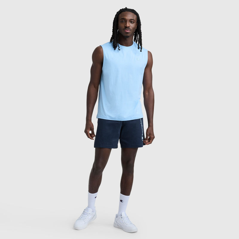 Man standing against a plain background, wearing a light blue sleeveless shirt, Champion 7″ Jersey Shorts, Script Logo in navy, white socks, and white sneakers. He has long hair and a relaxed expression.