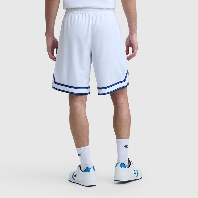 A person stands facing away, wearing Champion’s 10" Gridiron Reversible Short with Script Logo (blue trim), a white shirt, white crew socks, and white sneakers with blue accents on a plain light background.