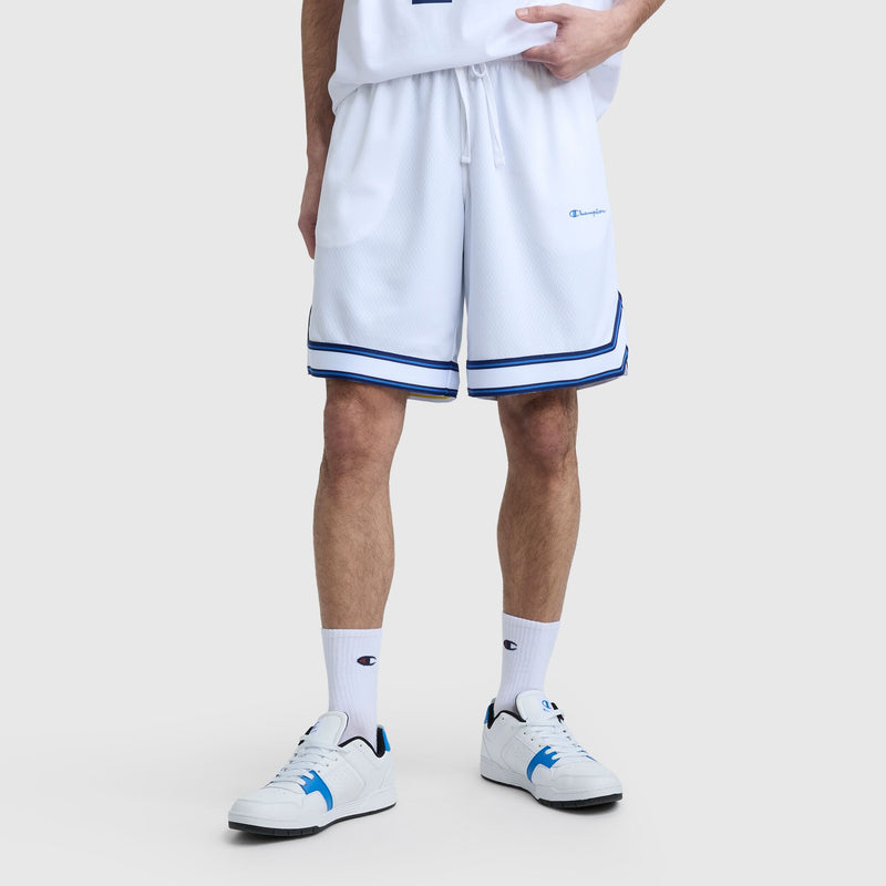 Someone stands against a light gray background wearing Champion's 10" Gridiron Reversible Short with blue trim, a matching white shirt, white crew socks, and white sneakers featuring blue accents.