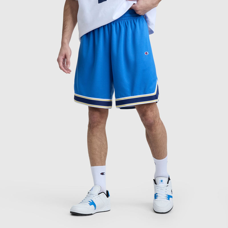 A person wears blue Champion 10" Gridiron Reversible Shorts with a script logo, white socks, and white sneakers with blue accents, standing against a plain light background. Their upper body is partially visible.