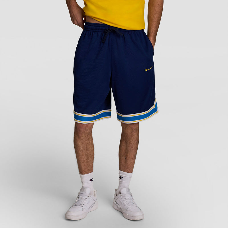 A person wearing a yellow shirt, Champion 10" Gridiron Reversible Short, Script Logo—navy reversible athletic shorts with yellow and white stripes—plus white socks and sneakers stands with hands in pockets against a plain backdrop.