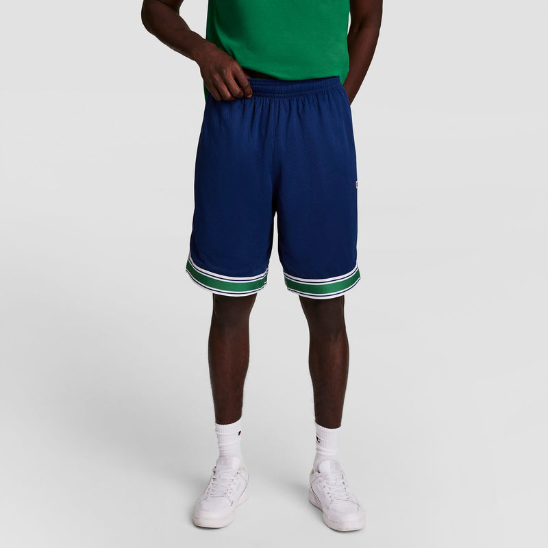 A person wearing a green shirt, blue athletic shorts with white and green trim, white socks, and white sneakers stands against a plain light gray background. The photo is cropped above the shoulders.