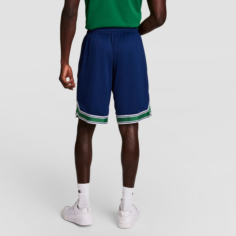 A person wearing a green shirt, navy blue athletic shorts with white and green trim, white socks, and white sneakers is standing with their back to the camera against a plain light background.
