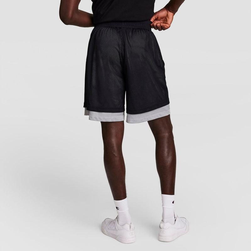 A person stands with their back to the camera, wearing black and gray mesh athletic shorts, a black shirt, white socks, and white sneakers against a plain light background.