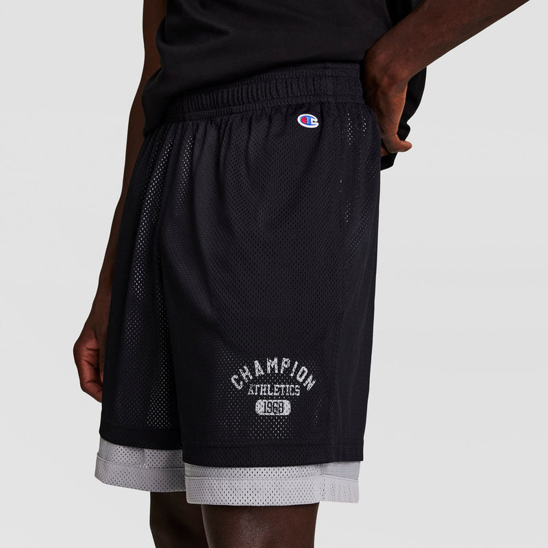 A person wearing black Champion athletic mesh shorts with a white Champion logo and ATHLETICS 1919 text near the hem, and a black shirt, standing against a plain light background.