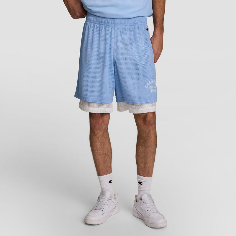 A person stands against a plain light background wearing Champion's 9" Gridiron Double Layer Short with C Logo in light blue over white athletic mesh shorts, paired with white socks and white sneakers.