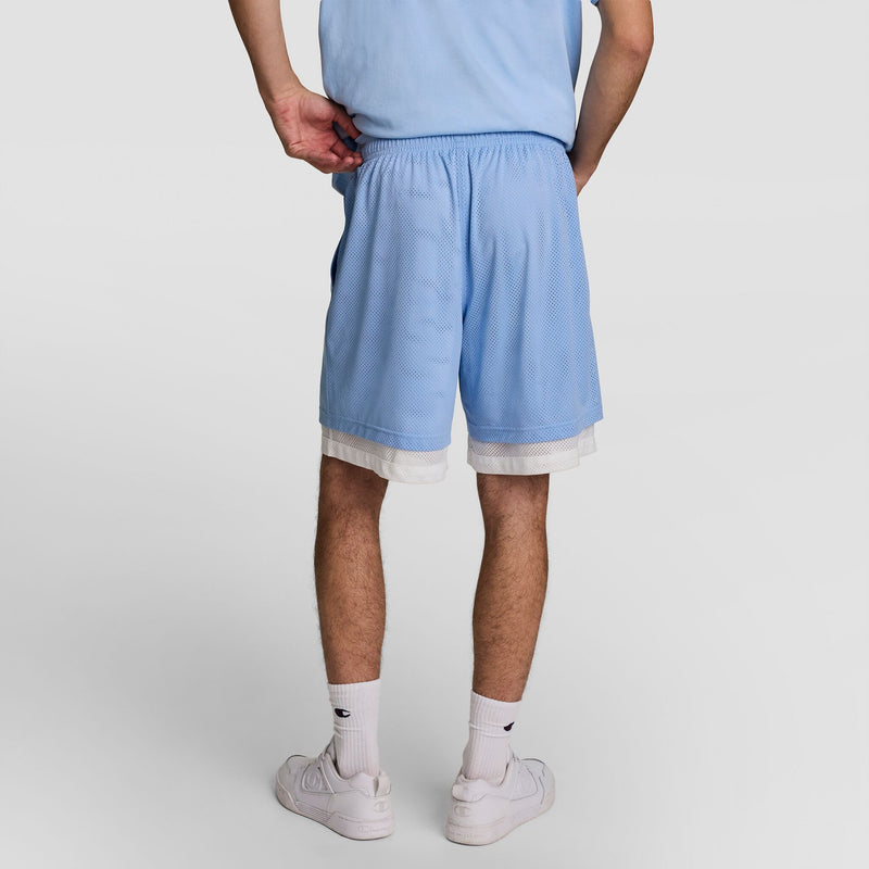 A person stands with their back to the camera, wearing Champion’s 9" Gridiron Double Layer Short, C Logo in light blue over white shorts, paired with a matching shirt, white crew socks, and white sneakers against a plain white background.