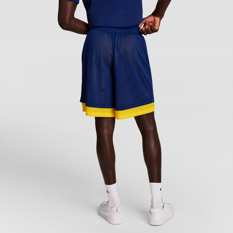 A person stands with their back to the camera, wearing a navy blue Champion Powerblend Full-Zip Hoodie, C Logo - Big & Tall over a navy basketball outfit with a yellow layer, paired with white socks and sneakers.